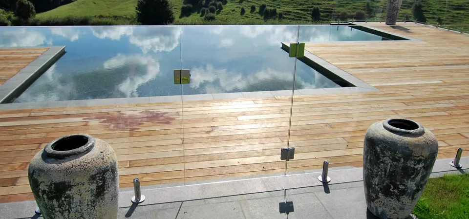 Glass pool fence beside a timber deck with sky reflected in the pool
