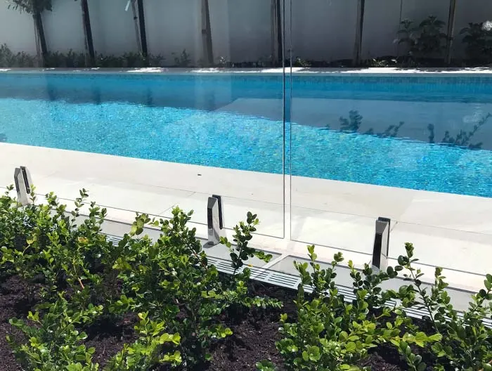 Close-up of glass pool fence panels fixed with spigots, with a hedge in front